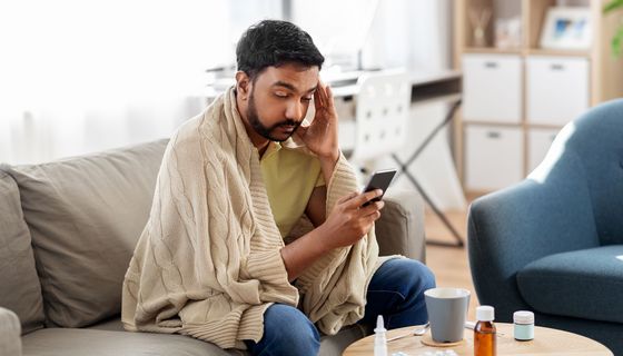 Ein Mann, der sich an die Schläfe fasst und ungesund aussieht, blickt eingehüllt in eine Decke auf sein Smartphone in seiner Hand. Vor ihm stehen auf einem kleinen Tisch eine Tasse sowie Medikamente.