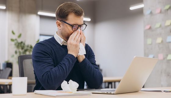 Junger Mann putzt sich im Büro die Nase. Vor ihm liegt bereits ein benutztes Taschentuch.